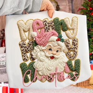 May include: White sweatshirt with an embroidered design featuring Santa Claus and the words "Merry Christmas." The text is in various colors, including cream, pink, green, and leopard print. The Santa Claus design includes a pink hat and a white beard.