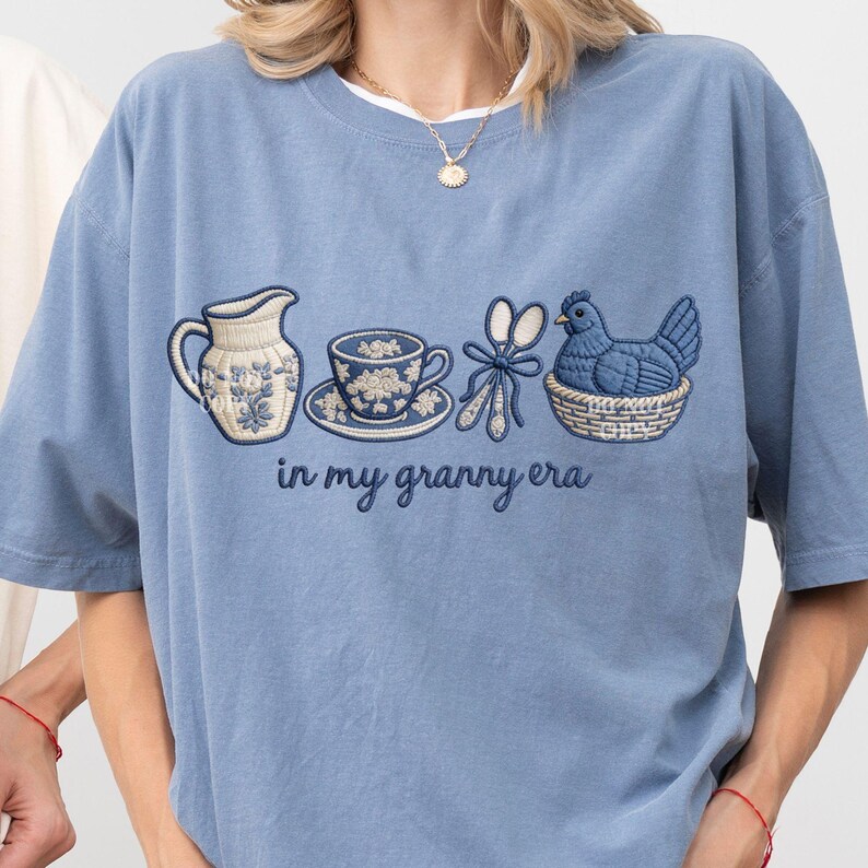 May include: A pale blue t-shirt with an embroidered design. The design includes a jug, teacup, scissors, and a hen in a basket. The words "in my granny era" are embroidered below. The shirt has a loose fit.