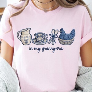 May include: Pale pink t-shirt with embroidered blue and white designs of a jug, teacup, scissors, and a hen in a basket. The text "in my granny era" is displayed below the images.
