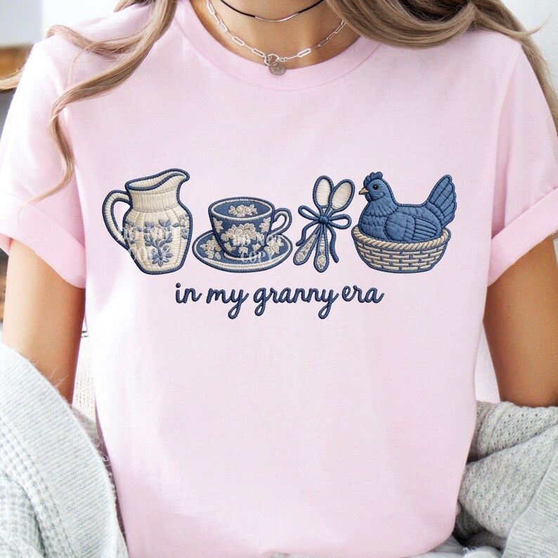 May include: Pale pink t-shirt with embroidered blue and white designs of a jug, teacup, scissors, and a hen in a basket. The text "in my granny era" is displayed below the images.