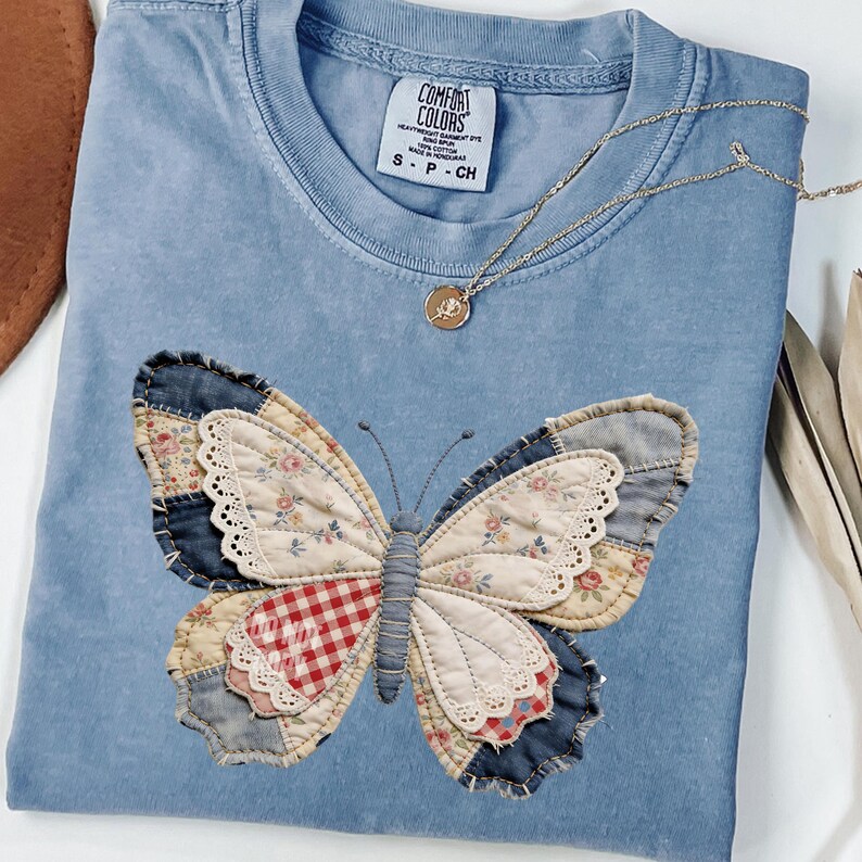 Pu&ograve; includere: Una maglietta Comfort Colors azzurra con un design a farfalla patchwork. Le ali della farfalla presentano motivi floreali, denim e a quadri. Una collana d'oro con un ciondolo poggia sulla maglietta. Taglia S-P-CH.