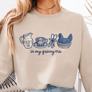 May include: Beige sweatshirt featuring embroidered blue and white floral motifs. The design includes a jug, teacup, scissors, and a hen in a basket, with the text "in my granny era".
