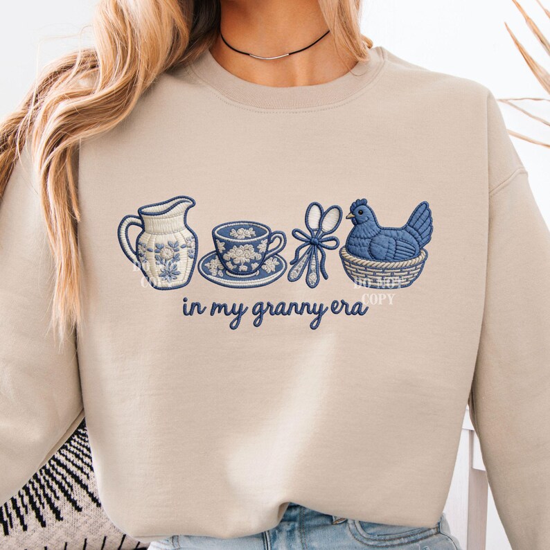 May include: Beige sweatshirt featuring embroidered blue and white floral motifs. The design includes a jug, teacup, scissors, and a hen in a basket, with the text "in my granny era".