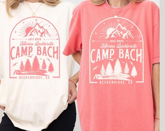 May include: Two white t-shirts with a pink graphic design. The design features a mountain range, a sun, trees, and the text "JULY 2025 Marias Bachlorette CAMP BACH BECKENRIDGE, CO".