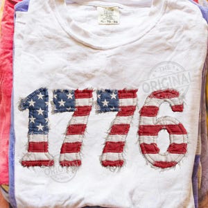 Pode incluir: Camiseta branca com o número "1776" aplicado na frente, apresentando o design da bandeira americana. Os números são em listras vermelhas e brancas com um quadrado azul e estrelas brancas. A camiseta está dobrada.