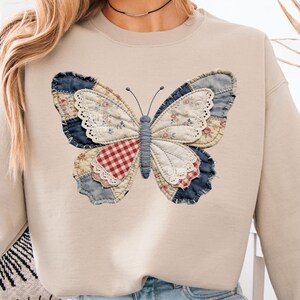 Pu&ograve; includere: Felpa beige con un grande disegno a farfalla. La farfalla &egrave; realizzata in tessuto patchwork con vari motivi, tra cui floreale, denim e scozzese rosso. La farfalla ha un corpo e antenne blu.