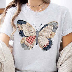 Pu&ograve; includere: T-shirt grigio chiaro con un'applicazione a farfalla di grandi dimensioni. La farfalla ha ali realizzate con patchwork di denim, tessuti floreali e a quadri. Il corpo della farfalla &egrave; di colore azzurro. La maglietta ha maniche corte.
