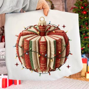 Może przedstawiać: Poduszka w kształcie bombki choinkowej z dzianinowym wzorem. Ozdoba wykonana jest z czerwonej, zielonej i kremowej tkaniny, przypominającej ułożone w stos książki. Widoczne są słowa "MERRY CHRISTMAS". Girlanda świetlna i gwiazdki otaczają ozdobę.