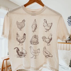 Może przedstawiać: Kremowa koszulka z szczegółowymi ilustracjami kurczaków w różnych pozach wykonanymi piórem i tuszem. Grafika zawiera kurczaki, koguty i kurę na gnieździe. Koszulka wisi na drewnianym wieszaku.