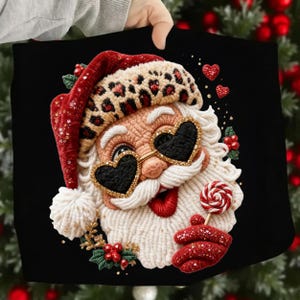 May include: A black Christmas-themed tote bag featuring a whimsical Santa Claus design. Santa wears heart-shaped sunglasses, a red hat with leopard print, and holds a lollipop. The design includes red hearts and berries.