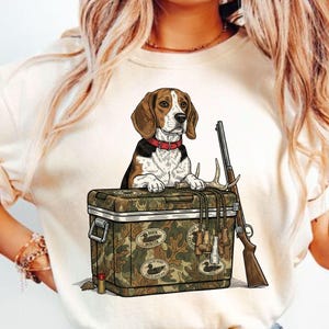 May include: Cream-colored t-shirt featuring a beagle dog wearing a red collar, standing behind a camouflage cooler. A rifle, antlers, and binoculars are also depicted. The cooler has "Duck Hunter" patches.