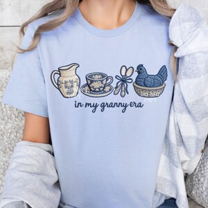 May include: Pale blue t-shirt with embroidered images of a jug, teacup, scissors, and a hen in a basket. The words "in my granny era" are printed below. The shirt is made of a soft, comfortable material.