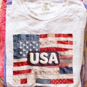 May include: White t-shirt with a patchwork American flag design. The flag incorporates blue, red, and white fabric pieces, with white stars and the word "USA" in a white, textured font. The shirt is XL-TG-EG.