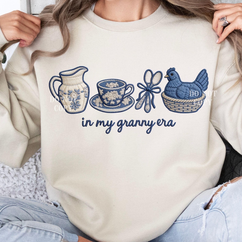 May include: Cream sweatshirt featuring embroidered blue and white floral motifs. The design includes a jug, teacup, scissors, and a hen in a basket, with the text "in my granny era" underneath.