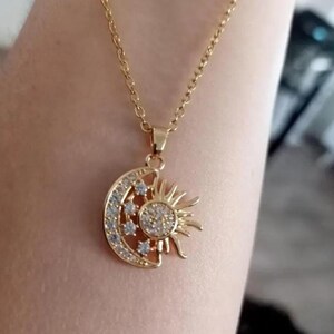 Peut inclure: Collier doré avec un pendentif représentant un soleil et une lune. Le soleil a une pierre centrale ronde et transparente et des rayons. La lune est en forme de croissant avec des pierres transparentes. La chaîne est délicate et dorée.