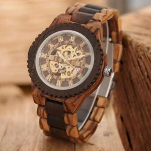 Peut inclure: Une montre-bracelet en bois avec un bracelet marron et noir. Le cadran de la montre présente des chiffres romains et un mouvement mécanique visible. La montre est en bois et a un aspect naturel et rustique.
