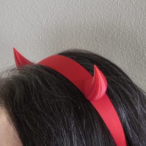 Könnte beinhalten: Ein rotes Teufelshorn-Stirnband. Das Stirnband ist einfarbig rot, mit zwei gebogenen Hörnern auf jeder Seite. Die Hörner sind ebenfalls rot und haben eine glatte, abgerundete Form. Das Stirnband wird auf dunklem Haar getragen.