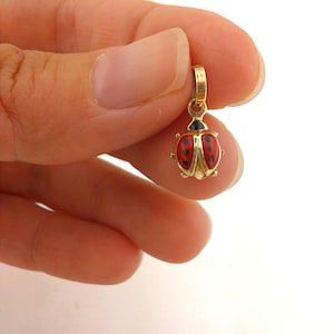 Puede incluir: Un dije de mariquita de color dorado con alas de esmalte rojo y lunares negros. El dije tiene un pequeño lazo en la parte superior para sujetarlo a un collar o pulsera. El cuerpo de la mariquita está detallado con detalles dorados.