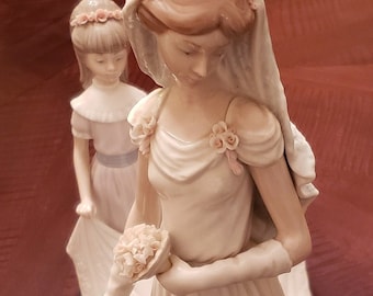 May include: A porcelain figurine of a bride and a flower girl. The bride is wearing a white gown with a veil and holding a bouquet of flowers. The flower girl is wearing a light blue dress with a white sash and a flower crown. The figurines are standing on a brown base.