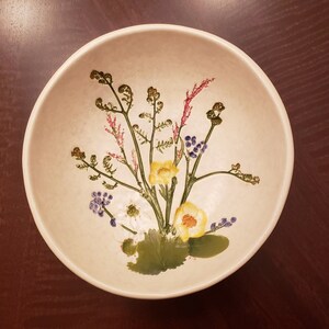 May include: A white ceramic bowl with a floral design. The bowl features a bouquet of yellow, white, and purple flowers with green leaves and stems.