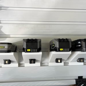 Puede incluir: Cuatro baterías de herramientas eléctricas en varios tonos de gris y negro se muestran en soportes de pared blancos. Las baterías están etiquetadas con nombres de marcas e información de voltaje. Los soportes están fijados a una pared de listones blancos.