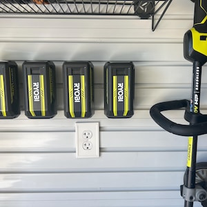 Può includere: Un gruppo di batterie nere e gialle per utensili elettrici Ryobi e un decespugliatore sono montati su una parete bianca. Le batterie sono etichettate con il logo Ryobi e sono disposte in fila. Il decespugliatore è nero e giallo.