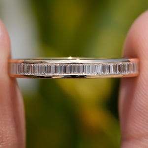 Puede incluir: Un anillo de banda de oro con una fila de diamantes de talla baguette. El anillo se sostiene entre dos dedos sobre un fondo borroso verde y amarillo.