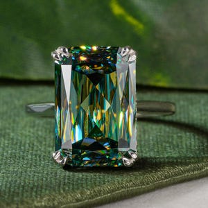 May include: A silver ring featuring a large, emerald-cut gemstone with a vibrant green and yellow hue. The ring is set against a backdrop of green fabric and a large green leaf, creating a luxurious and natural aesthetic.