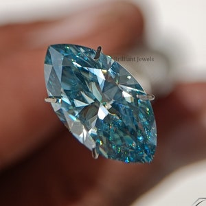 May include: A marquise cut blue diamond gemstone with a silver setting. The diamond is held by a pair of tweezers. The text "Brilliant Jewels" is visible in the image.