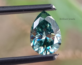 May include: A pear-shaped, teal green gemstone with facets, held by tweezers against a blurred green background. The text "Brilliant Jewels" is visible in the image.