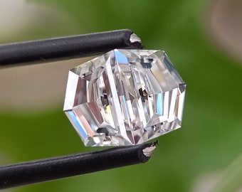 May include: A clear, octagonal-shaped gemstone with facets, held by tweezers against a green background.
