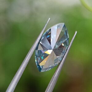 May include: A light blue marquise cut gemstone held by tweezers against a green background. The gemstone has a brilliant cut and facets that reflect light.