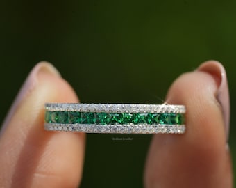 May include: A silver band ring with a row of emerald green square-shaped stones surrounded by smaller clear stones.