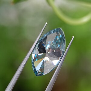 May include: A light blue marquise cut gemstone held by tweezers against a green background. The gemstone has a brilliant cut and facets that reflect light.