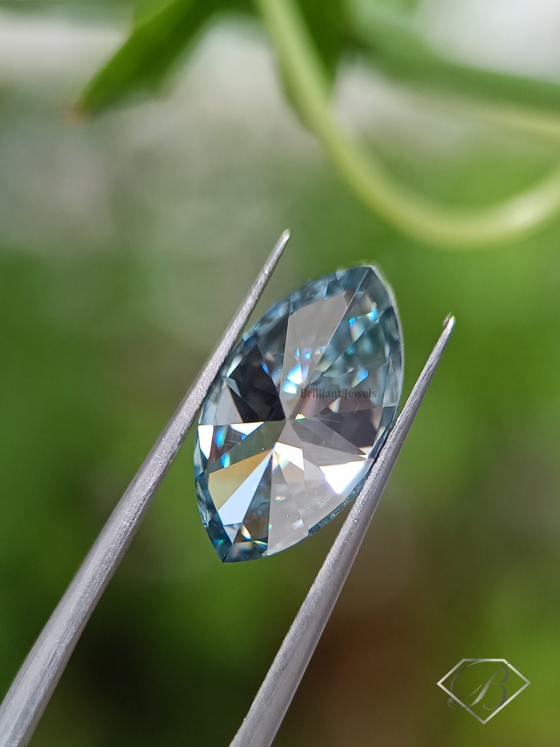 May include: A light blue marquise cut gemstone held by tweezers against a green background. The gemstone has a brilliant cut and facets that reflect light.