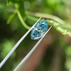 May include: A marquise-shaped blue moissanite gemstone held by tweezers against a green leafy background. The gemstone has a brilliant cut and sparkles with rainbow colours.  Brilliant Jewels is printed on the image.