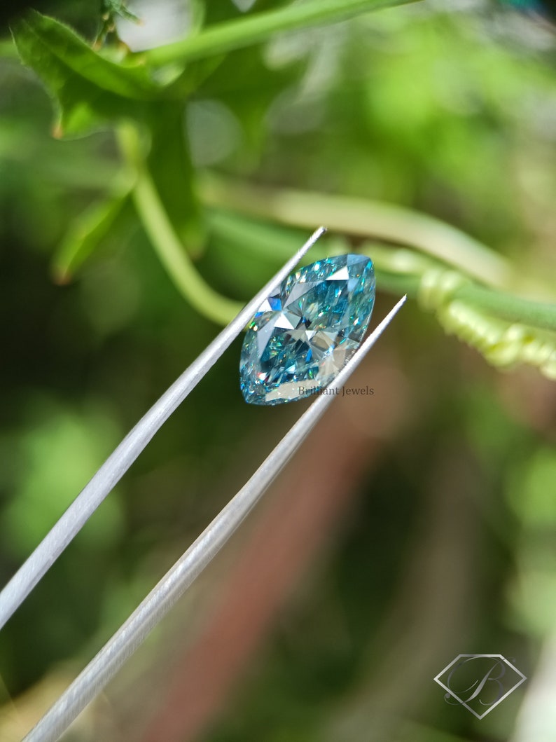 May include: A marquise-shaped blue moissanite gemstone held by tweezers against a green leafy background. The gemstone has a brilliant cut and sparkles with rainbow colours.  Brilliant Jewels is printed on the image.