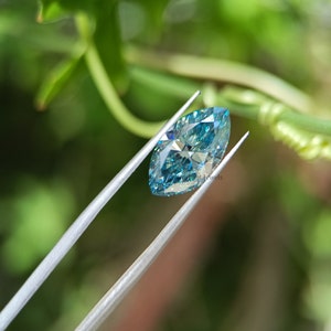 May include: A light blue marquise cut diamond gemstone held by tweezers against a green leafy background.