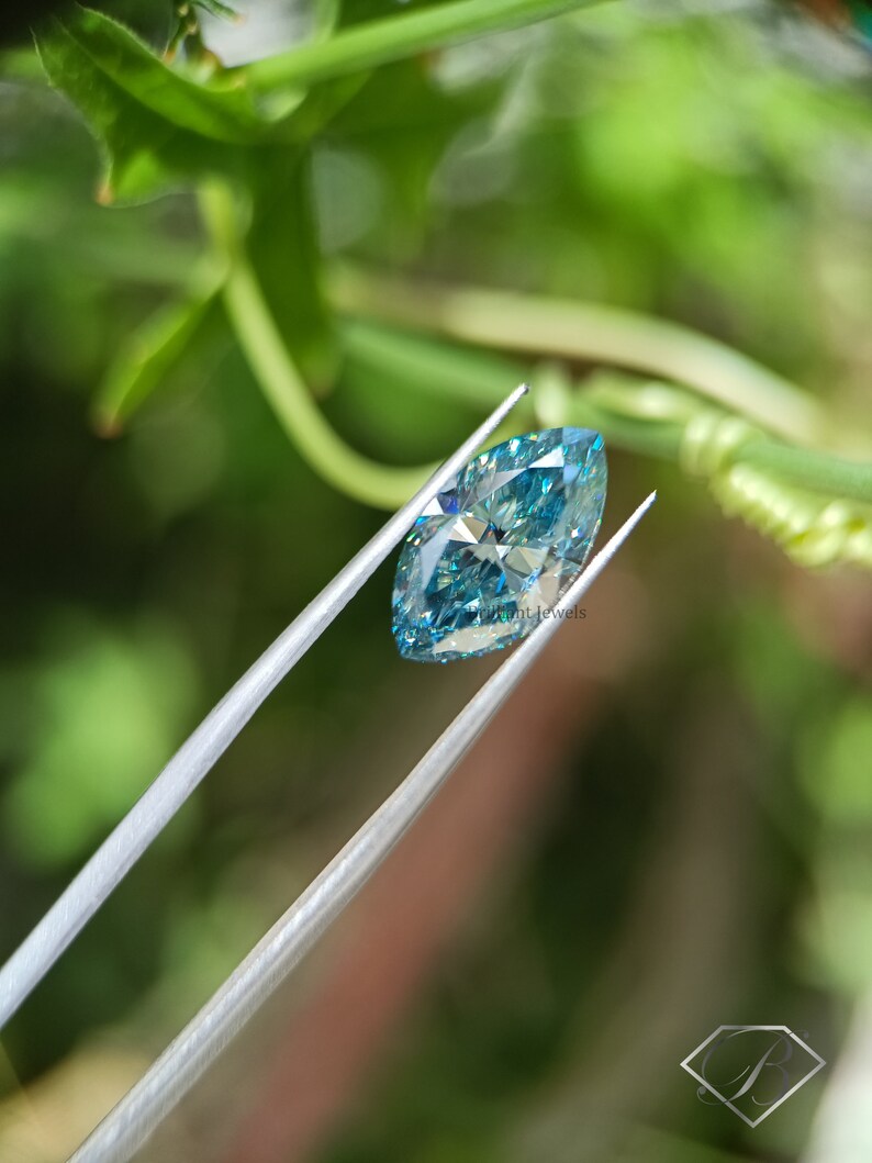 May include: A light blue marquise cut diamond gemstone held by tweezers against a green leafy background.