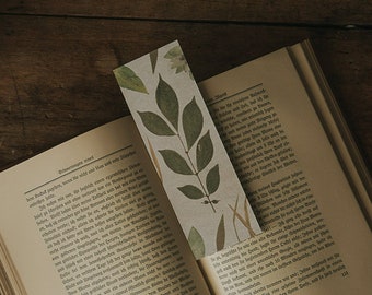 May include: A close-up of a book with a pressed leaf bookmark. The book is open to a page with text in German. The bookmark is a light brown color with a green leaf pressed into it. The leaf is a light green color and has a delicate, intricate pattern.
