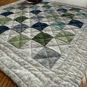 May include: A square quilted table topper featuring a grid of small fabric squares in shades of navy blue, forest green, sage, and light gray. The quilt has a white border with linear stitching and a textured pattern, topped with a glass candle jar.
