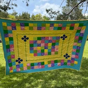 May include: A rectangular patchwork quilt featuring a central grid of multicolored squares surrounded by a solid yellow border. The quilt includes four dark blue flower appliqués at the corners and a bright turquoise outer border with yellow trim.