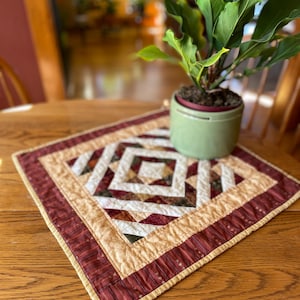 May include: A square quilted table topper featuring a geometric pattern in shades of burgundy, cream, and dark green. The center displays a diamond motif surrounded by borders of striped fabric and solid cream fabric, with a light green potted plant.