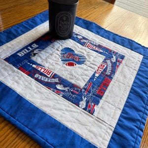May include: A square quilted fabric mat featuring a blue, white, and red Buffalo Bills pattern with a central heart-shaped Bills logo and a football graphic. A black cylindrical candle with the text SAND ALWO OD sits on the mat.