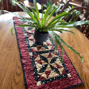 May include: A rectangular quilted table runner featuring a geometric star pattern in shades of deep red, cream, and dark green with a black border. A dark gray ceramic pot containing a green fern plant sits centered on the fabric runner.