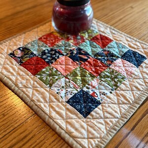 May include: A square quilted fabric mat featuring a patchwork design of various floral and geometric patterns in shades of red, blue, green, and white, bordered by a cream-colored diamond-stitched frame, topped with a glass candle jar.