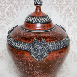Op de afbeelding: Een grote, rode keramische pot met een deksel en wolfshoofdaccenten. De pot heeft een gespikkelde glazuur en een zwarte en zilveren rand. Het deksel heeft een puntige bovenkant en is versierd met een zwart en zilveren patroon.