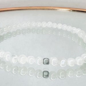 May include: A delicate white beaded bracelet with a small, square, silver-colored accent bead. The bracelet is made of small, round, white beads and is displayed on a reflective surface.