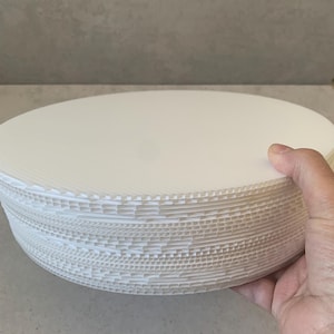 Op de afbeelding: Een stapel witte, ronde kartonnen taartborden. De borden worden in een hand gehouden, waardoor hun gegolfde randen zichtbaar worden. De borden zijn effen wit van kleur en lijken ongebruikt.