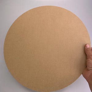 Op de afbeelding: Een grote, ronde cirkel van bruin karton. De rand van de cirkel wordt door een hand vastgehouden. Het karton heeft een gladde, matte afwerking. De achtergrond is een effen wit oppervlak.